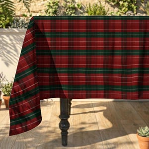 May include: A rectangular tablecloth featuring a classic plaid pattern with intersecting horizontal and vertical stripes in deep red, forest green, and thin white lines. The fabric drapes over the edge of a table, displaying the woven textile design.