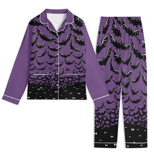 May include: A long-sleeve button-down pajama shirt and matching pants set in purple. The fabric features a repeating pattern of black bats flying against a purple background, transitioning to a solid black hem with white glowing eyes at the bottom.