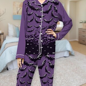 May include: A long-sleeve button-down pajama shirt and matching pants in purple, featuring a repeating pattern of black bats with white outlines. The shirt includes a black hem section with white dots and white piping along the collar and front.