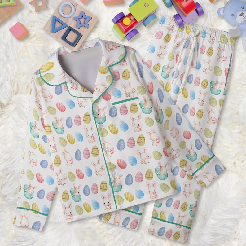 May include: White children's pyjama set featuring a pattern of Easter eggs and bunnies. The long-sleeved top and trousers have green trim. The pyjamas are made of a soft material.