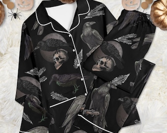 May include: A black pajama set featuring a long-sleeved button-down top and matching pants. The fabric displays a repeating pattern of black ravens, human skulls, and feathers against a dark background, accented with white piping along the edges.
