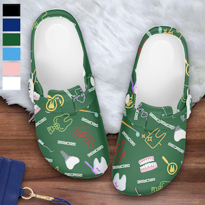 May include: A pair of green clogs with a white interior, featuring a pattern of dental tools, teeth, and the name "GEORGE" in white. The clogs are on a fluffy white surface.