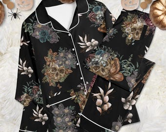 May include: A black long-sleeve button-down pajama shirt and matching pants set featuring a repeating print of human skulls, dark roses, and foliage. The shirt has a notched collar and white piping trim along the edges and cuffs.
