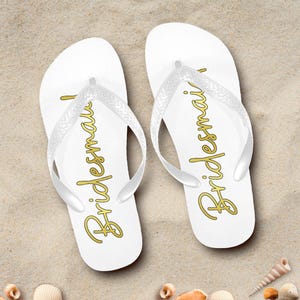 May include: White flip-flops with gold text that reads "Bridesmaid" are displayed on a sandy surface. The flip-flops have clear straps. Seashells are scattered around the flip-flops.