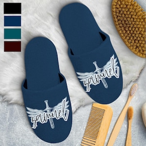 May include: A pair of navy blue slippers with a white graphic of a sword with wings and the name "Samuel" in a script font. The slippers are on a white, fluffy surface, with a wooden comb, toothbrushes, and a body brush in the background.