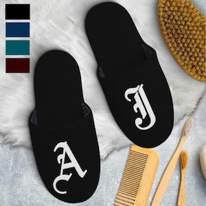 May include: Black slippers with white gothic-style letter designs. The slippers are displayed on a fluffy white surface, with a wooden comb, toothbrushes, and a brush in the background. A color palette of black, blue, teal, and burgundy is shown in the upper left corner.