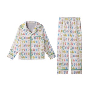 May include: A white Easter-themed pajama set featuring a button-down top and trousers. The set is patterned with colourful Easter eggs and bunny illustrations. The edges of the set are trimmed with green piping.