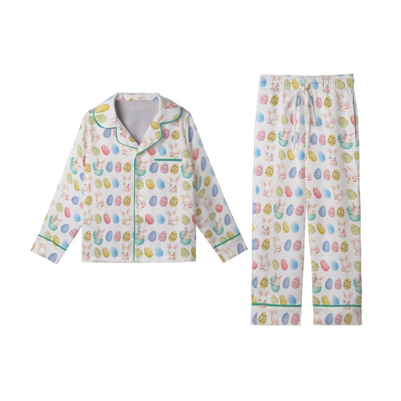 May include: A white Easter-themed pajama set featuring a button-down top and trousers. The set is patterned with colourful Easter eggs and bunny illustrations. The edges of the set are trimmed with green piping.