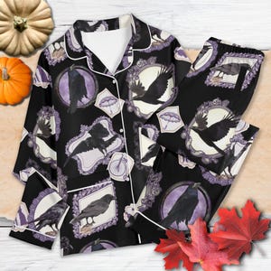 May include: Black pajama set with a repeating pattern of framed crows, umbrellas, and bicycles in purple and white. The set includes a button-down top and matching trousers, trimmed with white piping. The background is black.