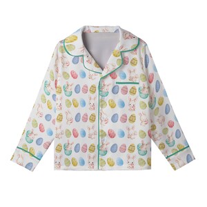 May include: White pajama top with an Easter pattern. The long-sleeved top features a repeating design of pastel-coloured Easter eggs and bunnies. The collar and trim are accented with a thin green border. The pajama top has a button-down front.