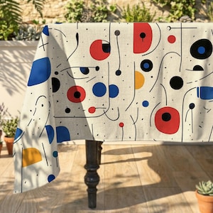Könnte beinhalten: Eine Tischdecke mit cremeweißem Untergrund und einem sich wiederholenden Muster aus schwarzen Linien, Punkten und ausgefüllten geometrischen Formen in Rot, Blau und Gelb. Das Design zeigt verschiedene Kreise, Halbkreise und geschwungene Linien.