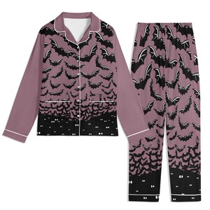 May include: A two-piece pajama set featuring a long-sleeved button-down shirt and matching pants. The fabric is a dusty mauve color with a repeating pattern of black bats. The bottom hem of the shirt and pants features a solid black border with white eyes.
