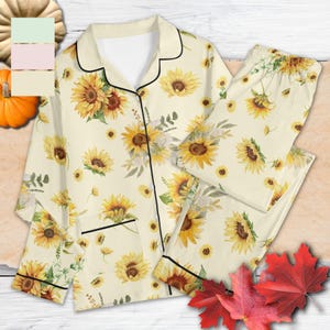 Könnte beinhalten: Zweiteiliger Schlafanzug in Creme mit Sonnenblumenmuster. Das langärmlige Oberteil hat schwarze Paspeln und eine Knopfleiste. Die passende Hose hat einen elastischen Bund. Das Set ist mit gelben Sonnenblumen verziert.