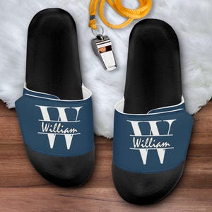 May include: A pair of black slide sandals with a navy blue strap featuring a large white letter W and the name William printed in white script. A yellow whistle on a matching yellow lanyard is positioned between the two sandals.