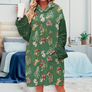 Op de afbeelding: Een groene hooded blanket met een patroon van rode, witte en bruine paddenstoelen. De deken heeft een grote voorzak en lange mouwen. De achtergrond is een slaapkamer.