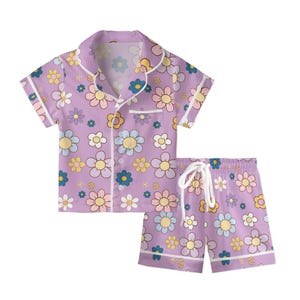 Puede incluir: Un conjunto de pijama lila con un estampado floral en tonos amarillos, azules y rosas. La parte superior de manga corta tiene botones y un bolsillo en el pecho. Los pantalones cortos a juego tienen cintura con cord&oacute;n y ribete blanco.