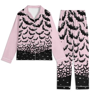 May include: A two-piece pajama set featuring a long-sleeved button-down shirt and matching pants. The fabric is light pink with a repeating black bat pattern that transitions into a solid black hemline featuring small white glowing eyes.