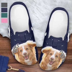 May include: Navy blue clogs featuring a smiling dog face with the name "Roger" printed on the side. The clogs have a white interior and a strap. The background is a fluffy white surface.