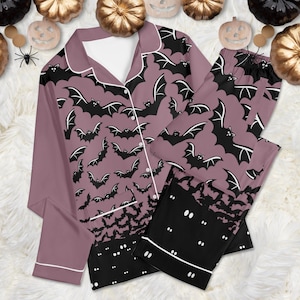 May include: A long-sleeved button-down pajama top and matching pants set in a dusty mauve color. The fabric features a repeating pattern of black bats with white wing details. The bottom hem of the shirt and pants features a solid black border.