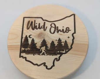 May include: A wooden round coaster with a laser-engraved design of the state of Ohio. The design includes a silhouette of a Bigfoot walking through a forest with trees and stars. The text "Wild Ohio" is engraved above the state outline.