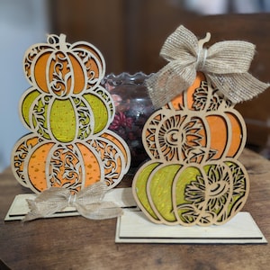 Op de afbeelding: Twee houten pompoenversieringen met oranje en groene lagen, ingewikkelde uitgesneden ontwerpen en jute strikken. De pompoenen zijn gestapeld en staan op rechthoekige voetstukken, perfect voor herfstdecoratie.