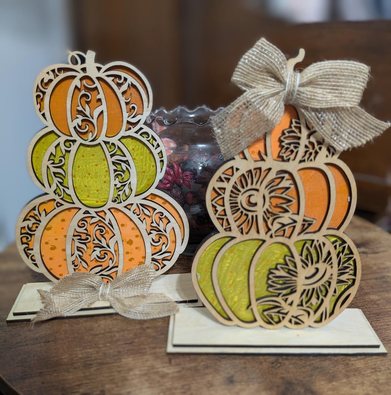 Op de afbeelding: Twee houten pompoenversieringen met oranje en groene lagen, ingewikkelde uitgesneden ontwerpen en jute strikken. De pompoenen zijn gestapeld en staan op rechthoekige voetstukken, perfect voor herfstdecoratie.