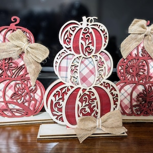 Op de afbeelding: Drie gestapelde houten pompoen decoraties met rood-witte ruit- en bloempatronen. Elke pompoen heeft een jute strik. De pompoenen staan op houten voetstukken, perfect voor herfstdecoratie.