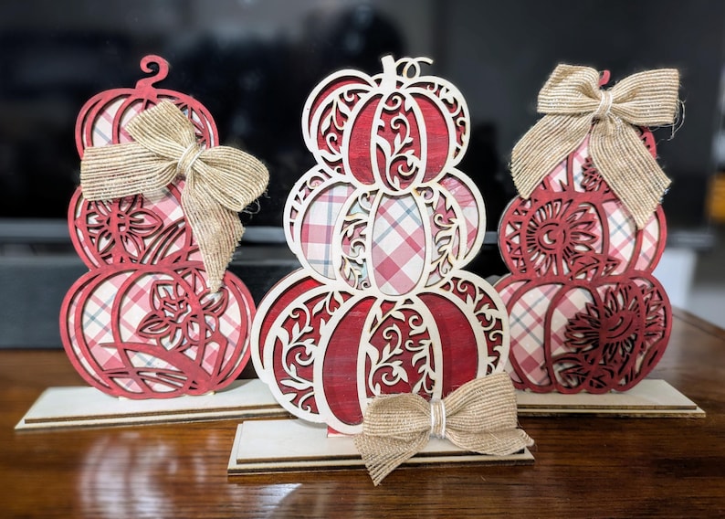 Op de afbeelding: Drie gestapelde houten pompoen decoraties met rood-witte ruit- en bloempatronen. Elke pompoen heeft een jute strik. De pompoenen staan op houten voetstukken, perfect voor herfstdecoratie.