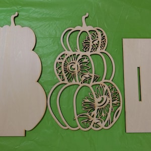 Op de afbeelding: Ongemaakte houten pompoenuitsparingen voor herfstdecoratie. De set bevat een effen pompoenvorm, een pompoen met zonnebloemuitsparingen en twee rechthoekige steunstukken. De pompoenen zijn gestapeld en hebben stelen.