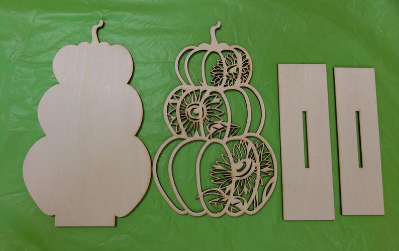Op de afbeelding: Ongemaakte houten pompoenuitsparingen voor herfstdecoratie. De set bevat een effen pompoenvorm, een pompoen met zonnebloemuitsparingen en twee rechthoekige steunstukken. De pompoenen zijn gestapeld en hebben stelen.