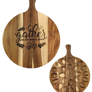 May include: Two wooden serving boards. One is round with a handle and the word "gather" engraved with the phrase "here with grateful hearts". The other board is round with a handle and has 19 oval indentations.