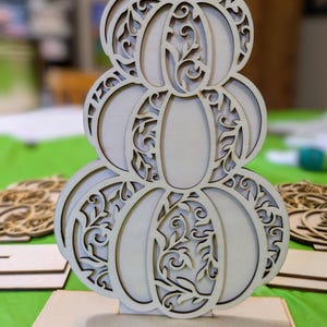 Op de afbeelding: Een houten pompoendecoratie met een gestapeld ontwerp. De pompoen is gemaakt van lichtgekleurd hout en heeft ingewikkelde uitgesneden patronen van wijnranken en bladeren. Hij staat op een rechthoekige houten basis, perfect voor herfstdecoratie.
