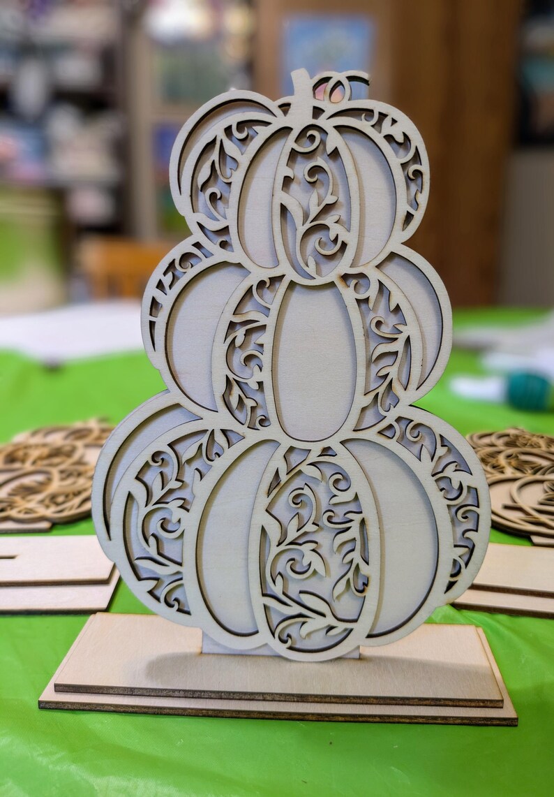 Op de afbeelding: Een houten pompoendecoratie met een gestapeld ontwerp. De pompoen is gemaakt van lichtgekleurd hout en heeft ingewikkelde uitgesneden patronen van wijnranken en bladeren. Hij staat op een rechthoekige houten basis, perfect voor herfstdecoratie.