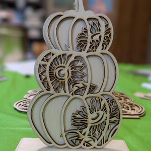 Op de afbeelding: Een houten pompoenvormige decoratie met drie gestapelde pompoenen. Elke pompoen heeft een uitgesneden zonnebloemontwerp. De decoratie is gemaakt van lichtgekleurd hout en staat op een rechthoekige basis. Perfect voor herfstdecoratie.