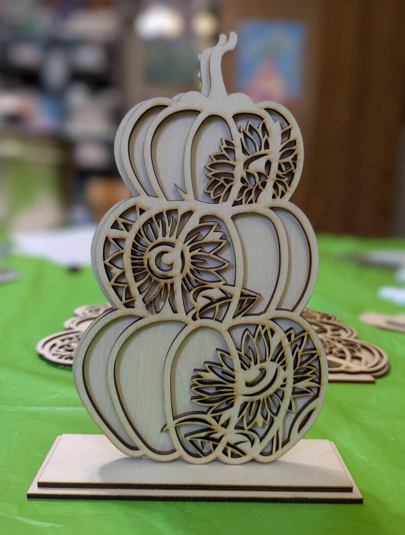 Op de afbeelding: Een houten pompoenvormige decoratie met drie gestapelde pompoenen. Elke pompoen heeft een uitgesneden zonnebloemontwerp. De decoratie is gemaakt van lichtgekleurd hout en staat op een rechthoekige basis. Perfect voor herfstdecoratie.