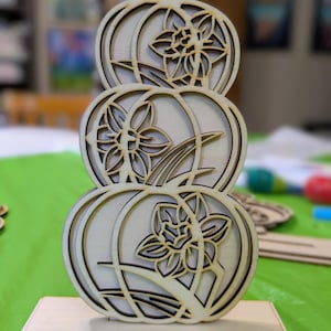 Op de afbeelding: Een houten decoratiestuk met drie gestapelde pompoenvormen, elk met een uitgesneden narcisontwerp. Het stuk is gemaakt van lichtgekleurd hout en staat op een rechthoekige basis. Het ontwerp is ingewikkeld en gedetailleerd.