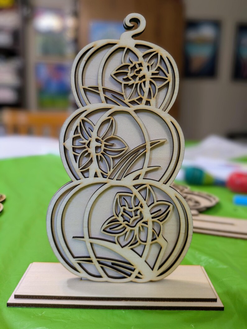 Op de afbeelding: Een houten decoratiestuk met drie gestapelde pompoenvormen, elk met een uitgesneden narcisontwerp. Het stuk is gemaakt van lichtgekleurd hout en staat op een rechthoekige basis. Het ontwerp is ingewikkeld en gedetailleerd.