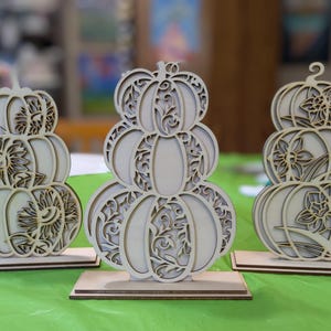 Op de afbeelding: Drie houten pompoenversieringen met ingewikkelde uitgesneden ontwerpen. Elke pompoenstapel heeft een ander bloemen- of zonnepatroon. De pompoenen worden tentoongesteld op een groen oppervlak, perfect voor herfstdecoratie.