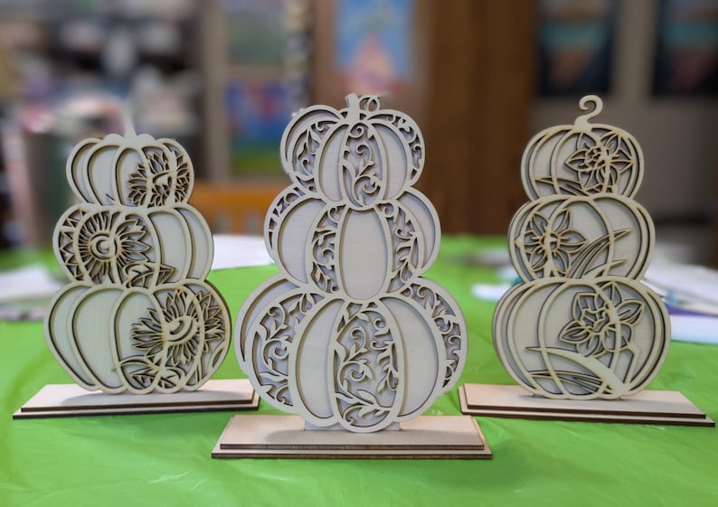 Op de afbeelding: Drie houten pompoenversieringen met ingewikkelde uitgesneden ontwerpen. Elke pompoenstapel heeft een ander bloemen- of zonnepatroon. De pompoenen worden tentoongesteld op een groen oppervlak, perfect voor herfstdecoratie.