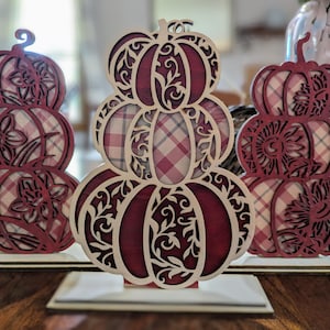Op de afbeelding: Drie decoratieve houten pompoen uitsparingen. Elke pompoen is gestapeld en heeft ingewikkelde bloemen- en wervelpatronen. De pompoenen hebben een bordeauxrood en cr&egrave;mekleurig ruitpatroon en worden tentoongesteld op kleine houten voetstukken.