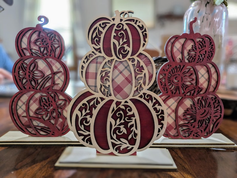 Op de afbeelding: Drie decoratieve houten pompoen uitsparingen. Elke pompoen is gestapeld en heeft ingewikkelde bloemen- en wervelpatronen. De pompoenen hebben een bordeauxrood en cr&egrave;mekleurig ruitpatroon en worden tentoongesteld op kleine houten voetstukken.
