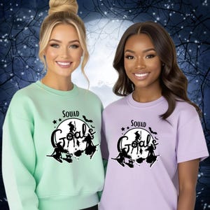 Könnte beinhalten: Zwei Frauen tragen passende Halloween-Oberteile. Eines ist ein hellgrünes Sweatshirt, das andere ein lavendelfarbenes T-Shirt. Beide haben eine schwarze Grafik mit "Squad Goals" und Hexensilhouetten auf Besen. Der Hintergrund ist eine gruselige Nachtszene.