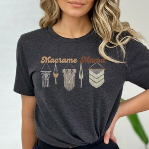 May include: A dark gray t-shirt with the text "Macrame Mama" in a retro font. The shirt features a design of five macrame wall hangings in various styles.