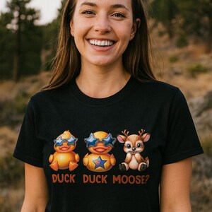 May include: A black t-shirt featuring three cartoon characters: two ducks wearing sunglasses and a deer. The text "DUCK DUCK MOOSE?" is printed below the characters. The person wearing the shirt is smiling.