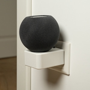 May include: A black, round smart speaker with a mesh grille is mounted on a white wall outlet.