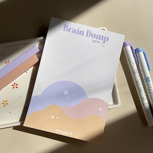 May include: A white notepad with the text "Brain Dump here" in purple lettering. The notepad has a pastel blue, pink, and yellow illustration of clouds and stars. The text "on my days off" is at the bottom of the notepad.