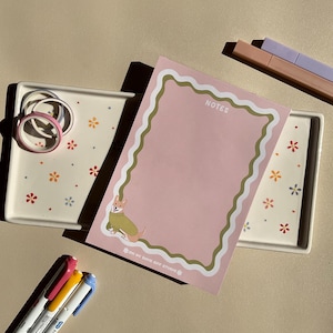 May include: A pink notepad with a green wavy border and the word "NOTES" is displayed. A cartoon corgi wearing a green sweater is in the corner. The notepad is next to a tray with floral designs, washi tape, and colorful highlighters.