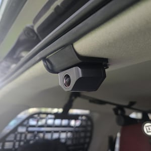 Może przedstawiać: Czarna, prostokątna kamera samochodowa jest zamontowana na wewnętrznym suficie pojazdu. Obiektyw kamery jest widoczny, a urządzenie jest przymocowane do czarnego wspornika. W tle widoczne jest wnętrze pojazdu.