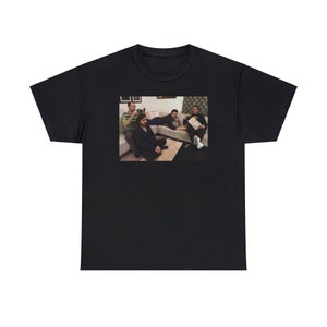 May include: A black t-shirt with a photo of four people sitting on a couch. The people are wearing casual clothing and are looking at the camera. The photo is in a rectangular frame and is centered on the shirt.
