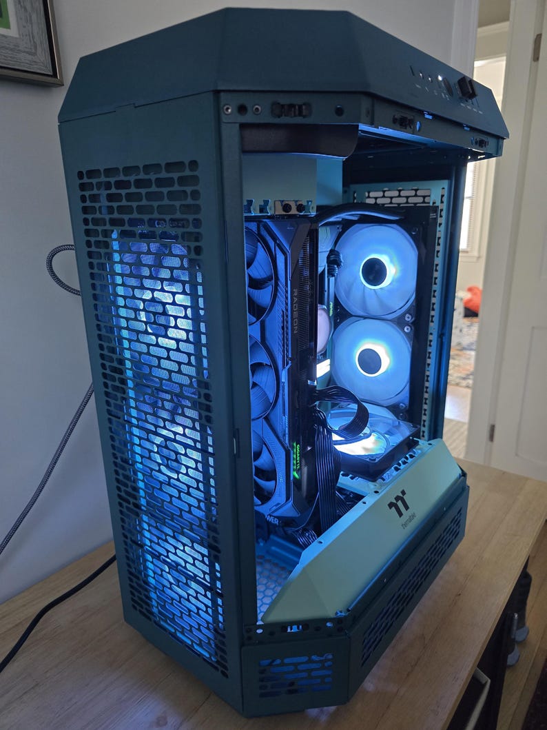 K&ouml;nnte beinhalten: Ein teal-farbenes Computergeh&auml;use mit einer Mesh-Frontplatte und internen Komponenten, die von blauen LED-Leuchten beleuchtet werden. Das Geh&auml;use hat ein einzigartiges, eckiges Design und verf&uuml;gt &uuml;ber mehrere L&uuml;fter.